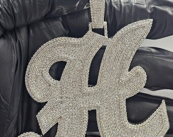 May include: A large, sparkling silver pendant in the shape of the letter "H". The pendant is encrusted with numerous small, clear gemstones, giving it a brilliant, eye-catching appearance. The bail is also covered in gemstones.