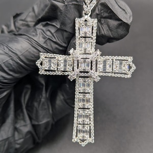 May include: A silver-colored pendant in the shape of a cross, encrusted with numerous rectangular-cut gemstones. The cross is held against a dark background. The pendant has a bail at the top for a chain.
