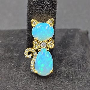 以下が含まれることがあります： 金色の猫のペンダント。淡いブルーオパールの体と頭が特徴です。猫の耳と尾には、小さな透明な宝石がちりばめられています。中央に透明な宝石が付いた蝶ネクタイの形で、頭と体を繋いでいます。