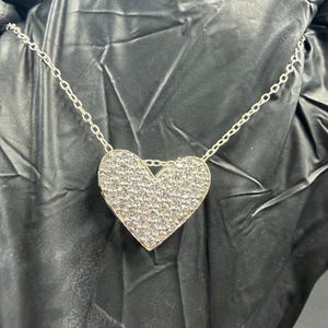 Peut inclure: Un collier argenté avec un pendentif en forme de cœur. Le pendentif est incrusté de petites pierres scintillantes, créant un effet chatoyant. La chaîne est délicate et est reliée au pendentif en forme de cœur.