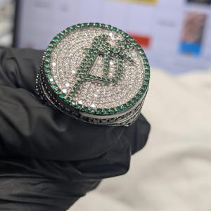 May include: A silver ring with a circular top, set with small, clear stones. The ring has a green letter "A" design in the centre, encircled by a border of green stones. The ring is held by a gloved hand.