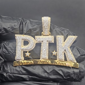 May include: Gold-toned pendant with the letters "PTK" encrusted with small, clear stones. The pendant has a curved base with the words "Prime Tyme Kennels" and two small star accents. The pendant is on a black surface.