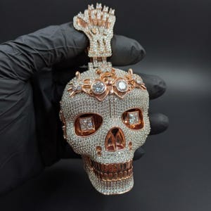 May include: A detailed pendant in the shape of a skull, encrusted with clear gemstones and rose gold accents. The skull features square-cut gems for eyes and a rose gold crown. The pendant is held against a black background.