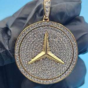 May include: A gold-colored pendant featuring a circular design with a three-pointed star emblem at the center. The pendant is encrusted with numerous small, clear gemstones, and has a loop at the top for a chain.