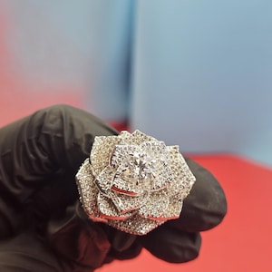 May include: A silver-toned ring in the shape of a rose, encrusted with numerous small, clear gemstones. The center of the rose features a larger, clear gemstone. The ring is held against a red and blue background.