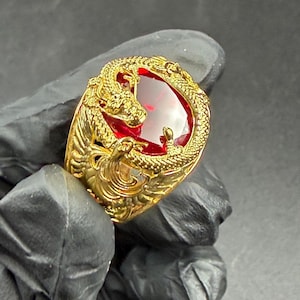 May include: A gold ring with a large, oval, red gemstone. The ring is decorated with a dragon design around the gemstone. The ring is held by a gloved hand against a dark background.