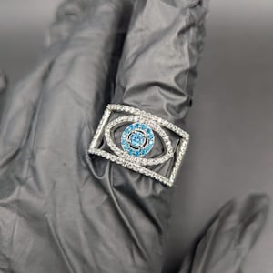 May include: A silver-toned ring featuring a rectangular design with a central eye motif. The eye is composed of a cluster of small, light blue gemstones surrounded by a border of clear, sparkling stones. The ring is set against a dark background.
