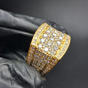 Puede incluir: Un anillo dorado con una banda ancha, densamente incrustado con numerosas piedras preciosas pequeñas y claras. El anillo se sostiene sobre un fondo oscuro, mostrando sus facetas brillantes y su diseño intrincado. La superficie del anillo es muy reflectante.