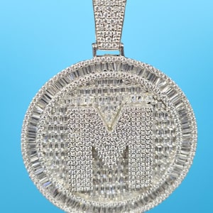 May include: A silver-colored pendant with the letter "M" encrusted with small, sparkling stones. The pendant has a circular design with a border of rectangular stones and a bail at the top for a chain.