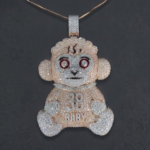Op de afbeelding: Een met diamanten bezette apenhanger aan een gouden ketting. De hanger heeft een cartoon apenontwerp met rode ogen en de tekst "38 BABY". De sieraden zijn geplaatst tegen een donkergrijze achtergrond.