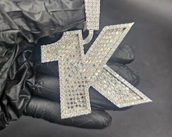 May include: A silver-toned pendant in the shape of "1K" encrusted with numerous small, sparkling stones. The pendant is held against a dark background, showcasing its reflective surface and intricate design. The pendant is likely a piece of jewelry.
