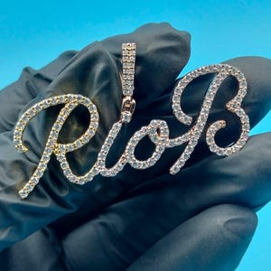 Puede incluir: Un colgante de oro y plata que deletrea "Rio B" en una fuente cursiva. Las letras están incrustadas con pequeñas piedras brillantes. El colgante se sostiene sobre un fondo negro.