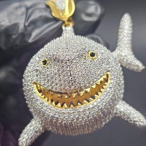 May include: A diamond-encrusted shark pendant with a gold-colored toothy grin and accents. The pendant features black eyes and a gold-colored bail. The shark's body is covered in small, sparkling stones.