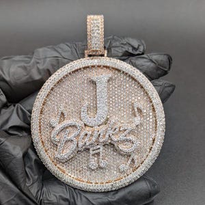 May include: A large, round pendant with a gold and silver design. The pendant features the letter "J" and the name "Banks" in a cursive font, all encrusted with small, sparkling stones. The pendant is held by a gold bail.