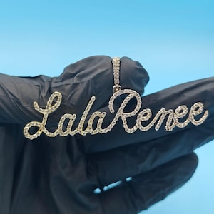 以下が含まれることがあります： 「Lala Renee」と筆記体で書かれた、輝く宝石がちりばめられたゴールドトーンのペンダント。ペンダントは、お揃いのベイルから吊り下げられています。背景は単色の水色です。