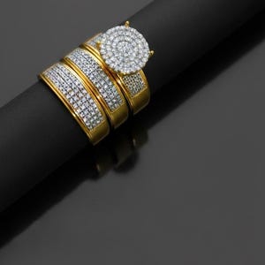 May include: A set of three gold-toned rings, each encrusted with small, sparkling clear stones. One ring features a large, circular cluster of stones. The rings are displayed on a black surface, creating a contrast that highlights the jewelry.