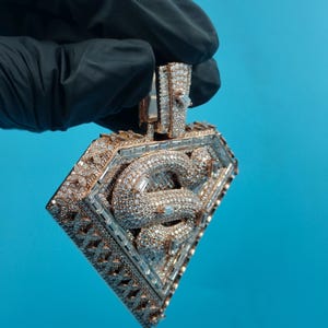 May include: A diamond-encrusted pendant in a diamond shape, held by a gloved hand. The pendant features a central design of intertwined snakes, surrounded by a frame of smaller diamonds. The metal has a gold tone.