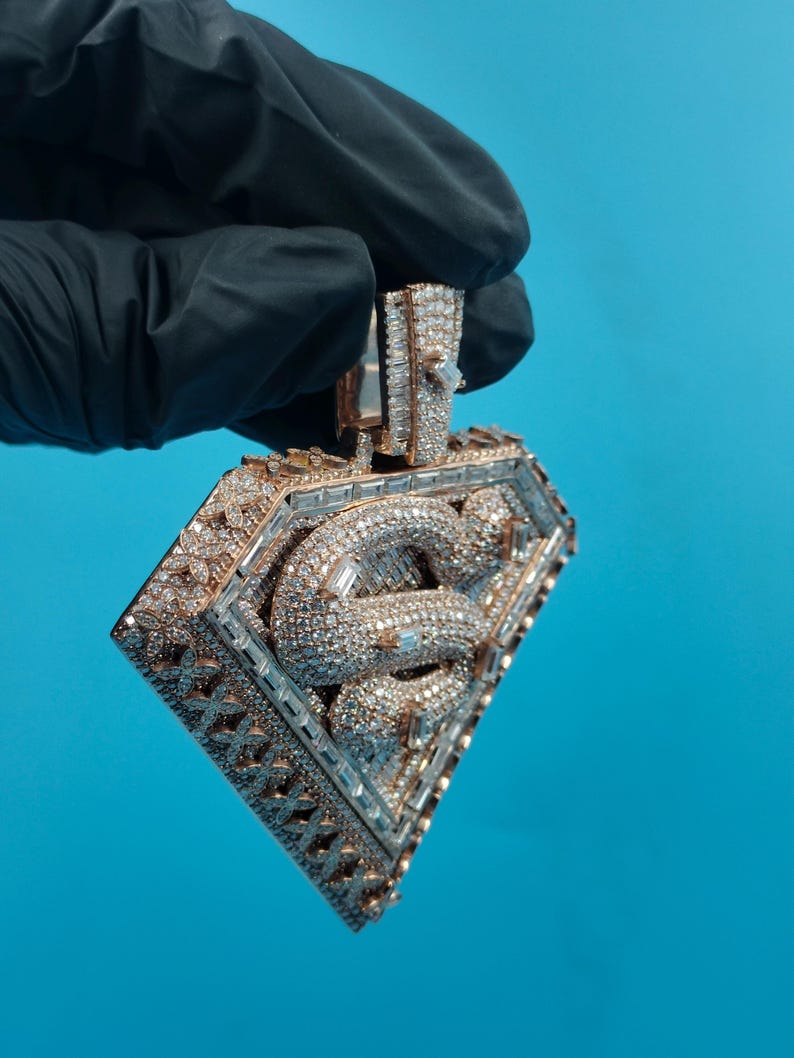 May include: A diamond-encrusted pendant in a diamond shape, held by a gloved hand. The pendant features a central design of intertwined snakes, surrounded by a frame of smaller diamonds. The metal has a gold tone.