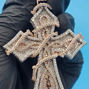 May include: A diamond-encrusted cross pendant with a rose gold and silver design. The pendant features a pink gemstone at the top and the word "JAZZE" in gold lettering. The cross is held by a gloved hand against a blue background.