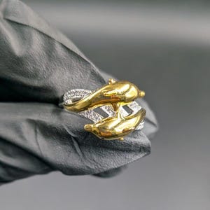 May include: A gold and silver ring featuring two dolphin figures. The dolphins are gold-colored, and the band is silver with small, clear stones. The ring is a unique piece of jewelry.
