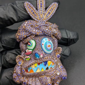 May include: A unique, ornate pendant featuring a whimsical face design. The pendant is encrusted with purple gemstones and gold accents. The face has colorful enamel details, including a blue and yellow mouth, and multi-colored eyes.