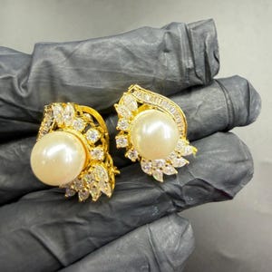 Peut inclure: Une paire de boucles d'oreilles dorées, chacune ornée d'une grosse perle ronde de couleur crème. Les boucles d'oreilles sont ornées de cristaux clairs et étincelants et de motifs complexes en forme de feuilles. Les boucles d'oreilles sont posées sur un fond sombre.