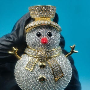 May include: A sparkling snowman pendant with a gold hat, scarf, and arms. The snowman's body is covered in small, clear stones, with black eyes and a red nose. The pendant is held against a blue background.