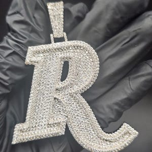 May include: A silver-colored pendant in the shape of the letter "R", encrusted with numerous small, sparkling stones. The pendant has a bail at the top for a chain. The background is a dark, solid color.