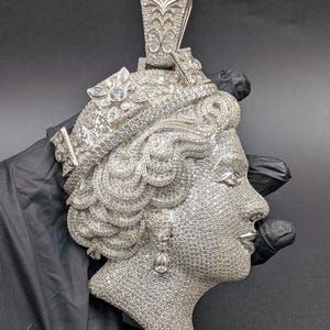Puede incluir: Un colgante plateado que representa el perfil de un rostro femenino, incrustado con numerosas piedras pequeñas y brillantes. El colgante tiene una fianza en forma de corazón y un pendiente en forma de lágrima. El diseño es muy detallado y ornamentado.