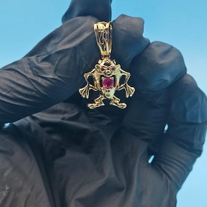 May include: A gold-colored pendant featuring the Tasmanian Devil cartoon character. The pendant has a red gemstone in the center and is suspended from a matching bail. The background is a solid light blue.