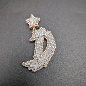 May include: A pendant featuring a large letter "D" and a star, both encrusted with small, sparkling crystals. The pendant has a gold-colored border. The star is attached to the top of the letter "D".