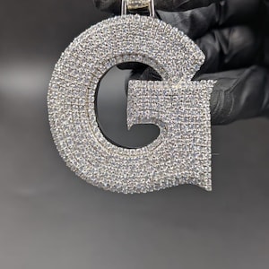 May include: A silver pendant in the form of the letter 'G', covered in many small, glittering stones. The pendant is held by a silver bail and is shown against a dark backdrop.
