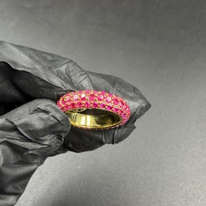 以下が含まれることがあります： 多数の小さな鮮やかなピンク色の宝石がちりばめられたゴールドリング。リングは暗い背景に対して保持され、ジュエリーの明るい色と複雑なデザインを際立たせています。リングのバンドは磨かれたゴールドです。
