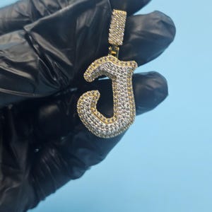Puede incluir: Un colgante dorado con forma de la letra "J", incrustado con pequeñas piedras blancas brillantes. El colgante se sostiene sobre un fondo azul claro. La argolla también está cubierta de piedras blancas.