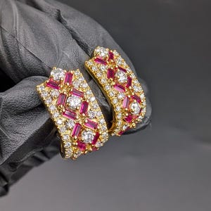 May include: A pair of gold earrings with a rectangular design. The earrings are set with small, round, and rectangular-cut white and red gemstones. The gemstones are closely set, creating a sparkling effect. The earrings are held against a dark background.