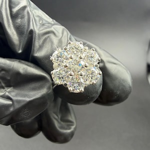 Op de afbeelding: Een clusterring met meerdere ronde, heldere edelstenen gerangschikt in een bloemmotief. De ring is gezet op een zilveren band, waarbij de edelstenen schitteren tegen een donkere achtergrond. De ring wordt vastgehouden door een gehandschoende hand.