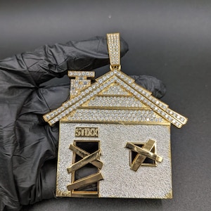 Puede incluir: Colgante de casa de oro y plata con techo y detalles con diamantes. La casa tiene ventanas tapiadas y la palabra "ST1XX" es visible. El colgante es una pieza de joyería única.