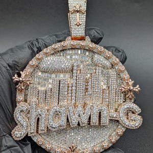 May include: A large, ornate pendant with the name "Shawn G" spelled out in sparkling, diamond-like stones. The pendant features a cityscape design and is set in a gold-colored metal with intricate detailing and cross-shaped accents.