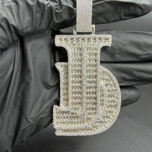 May include: A silver-colored pendant in the shape of the letter "JD", encrusted with numerous small, clear gemstones. The pendant has a rectangular bail at the top for a chain. The gemstones are set closely together, creating a sparkling effect.