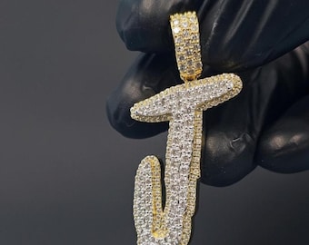 May include: A gold-toned pendant in the shape of the letter "J", encrusted with numerous small, sparkling stones. The pendant is held against a dark background, highlighting its bright, reflective surface. The letter is in a cursive style.