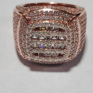 May include: Rose gold ring with a square, gem-set centre. The ring is covered in small, clear stones, with a band of smaller stones around the edges. The design is symmetrical and eye-catching, a statement piece.