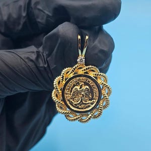 May include: Gold pendant featuring a Mexican coin design. The coin is set in a black background, surrounded by a twisted rope-like border. A small diamond is set at the top of the pendant.