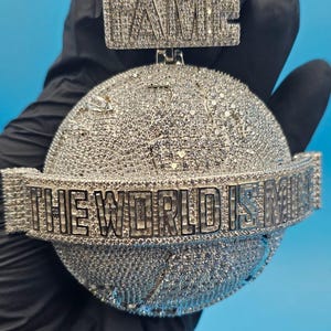 May include: A silver pendant with the word "FAME" at the top and a globe design. The phrase "THE WORLD IS MINE" is written around the globe. The pendant is covered in small, sparkling stones.