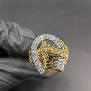 Puede incluir: Anillo dorado con un diseño de cabeza de caballo dentro de una herradura. El anillo tiene un fondo negro y está incrustado con piedras claras y brillantes. El anillo es una pieza llamativa, probablemente para un entusiasta de las carreras de caballos.