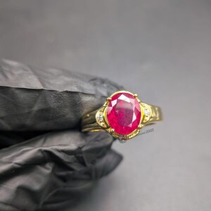 Puede incluir: Un anillo de oro con una gran piedra preciosa de color rojo vibrante y corte ovalado en un engaste de garras. La banda incluye pequeñas piedras de acento claras a ambos lados de la piedra central. El texto "gemmine2005" es visible en el anillo.