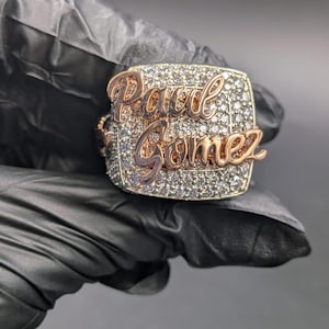 Puede incluir: Un anillo personalizado con el nombre "Paul Gomez" en escritura de oro rosa. La cara del anillo está incrustada con pequeñas piedras transparentes, engastadas en forma cuadrada con un borde de oro rosa. El anillo se muestra sobre un fondo negro.