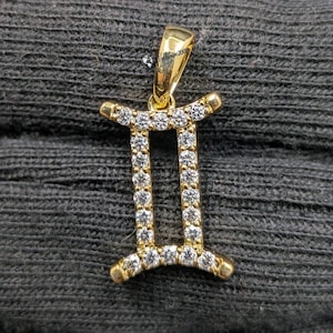 Peut inclure: Un pendentif doré en forme de symbole du zodiaque Gémeaux, composé de deux barres verticales reliées par des barres horizontales en haut et en bas. Toute la surface est incrustée de petites pierres claires et scintillantes. Une bélière est fixée au sommet.