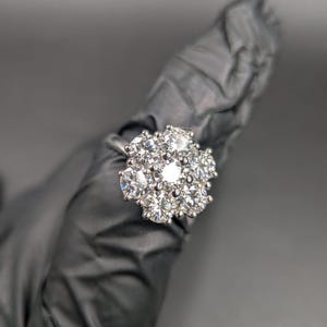 May include: A silver ring with a cluster of clear, sparkling stones. The stones are arranged in a floral pattern, creating a dazzling display. The ring is held against a dark background.