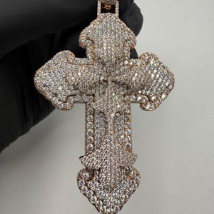 May include: A large, ornate pendant in the shape of a cross. The pendant is rose gold in color and encrusted with numerous small, sparkling stones. The cross design features intricate details and a bail for a chain.