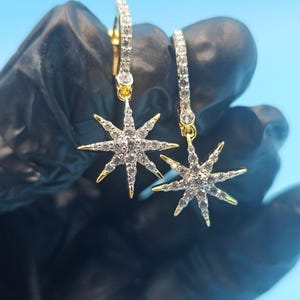 Peut inclure: Une paire de boucles d'oreilles en forme d'étoile dorée avec des cristaux clairs et étincelants. Les boucles d'oreilles ont un design à créoles et sont posées sur un fond bleu.
